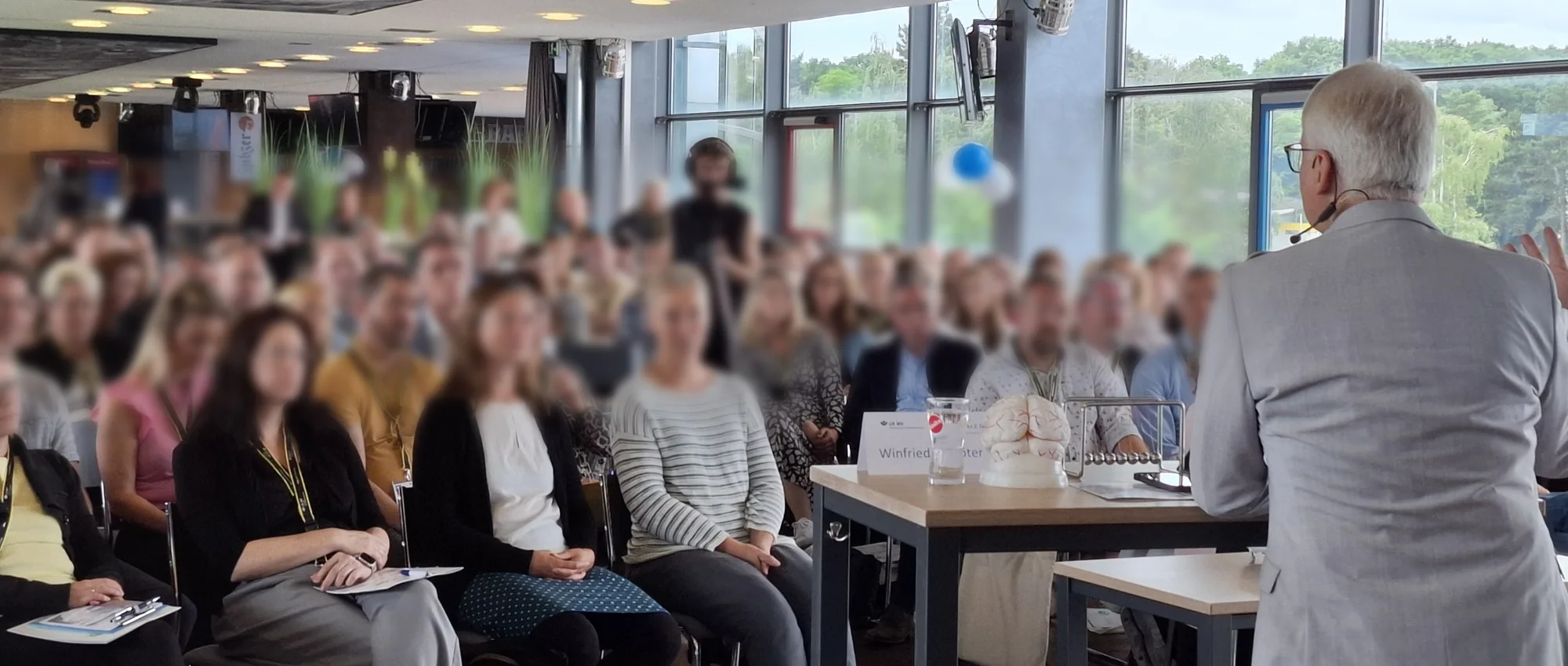 Winfried Schröter bei einem Vortrag vor vollbesetztem Saal, im Vordergrund sein Namensschild und ein Gehirnmodell